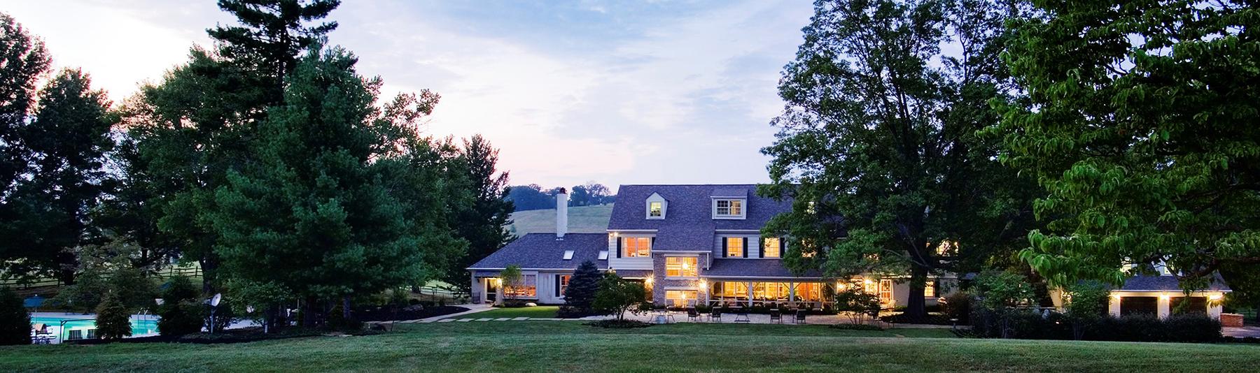 The back of the Estate House at dusk, its windows lit, with the pool and the lawn spread out across the grounds