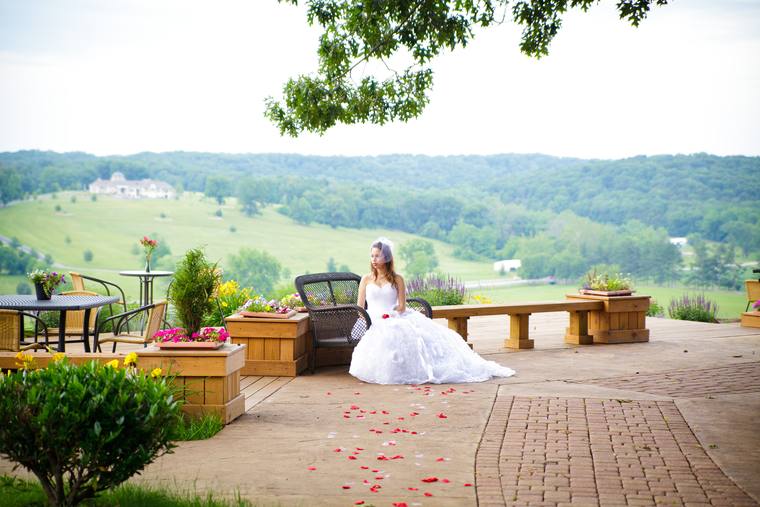 A bride seated on the hilltop terrace, rose petals scattered across the paving and the hills beyond