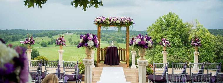 The hilltop dressed for a ceremony — a timber arbor hung with white and purple flowers, tall urn arrangements either side of the aisle, chairs facing the valley