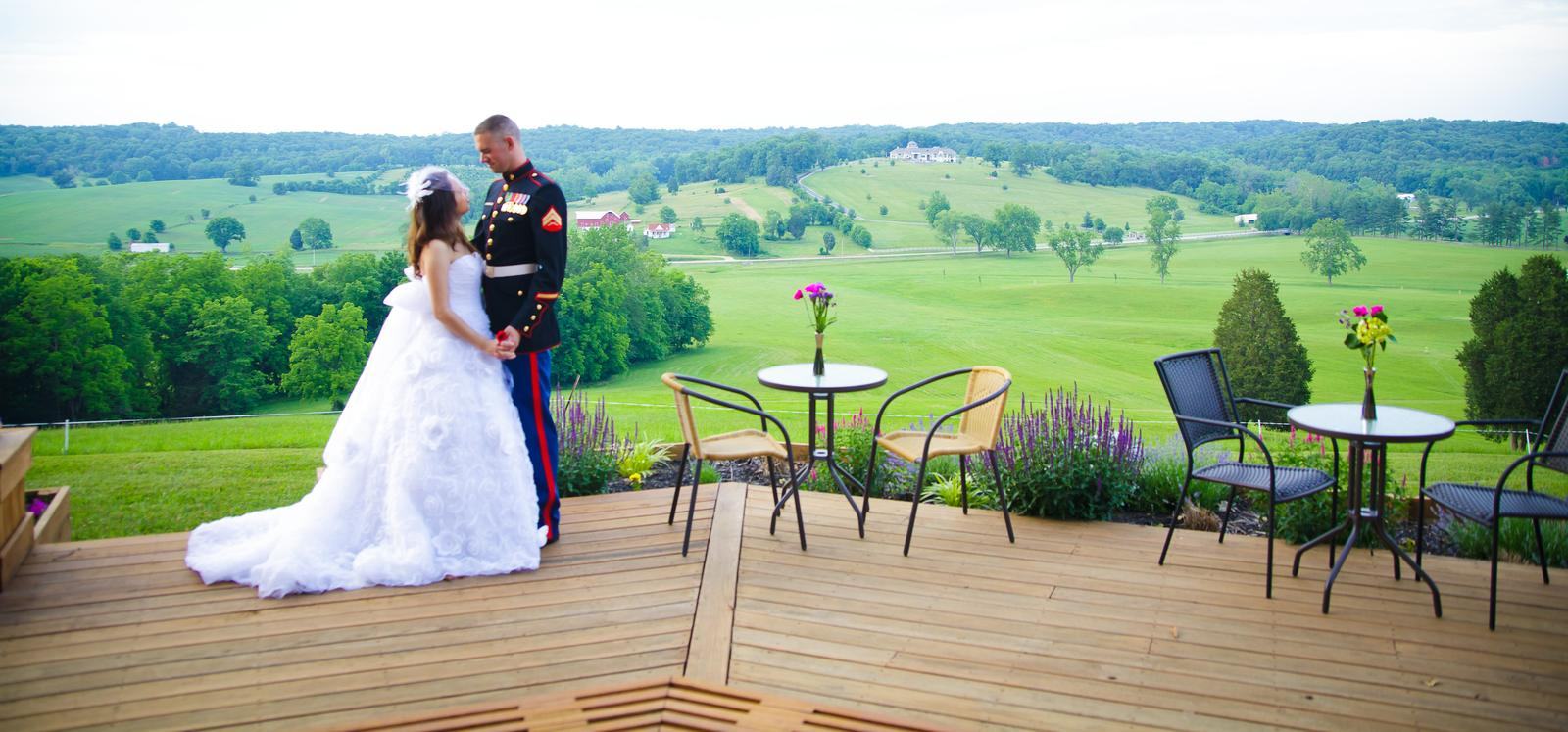 A bride and her groom in Marine dress blues together on the hilltop deck, café tables set beside them and the valley opening out beyond