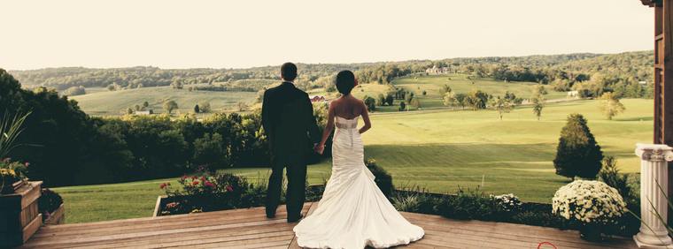A bride and groom standing hand in hand at the edge of the hilltop deck, looking out over the valley in warm evening light