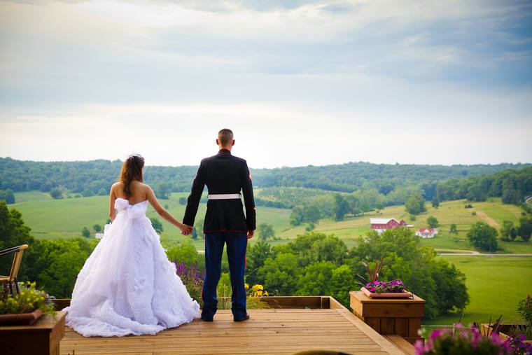 A bride and her groom in Marine dress blues, hand in hand at the edge of the hilltop deck, looking out across the valley